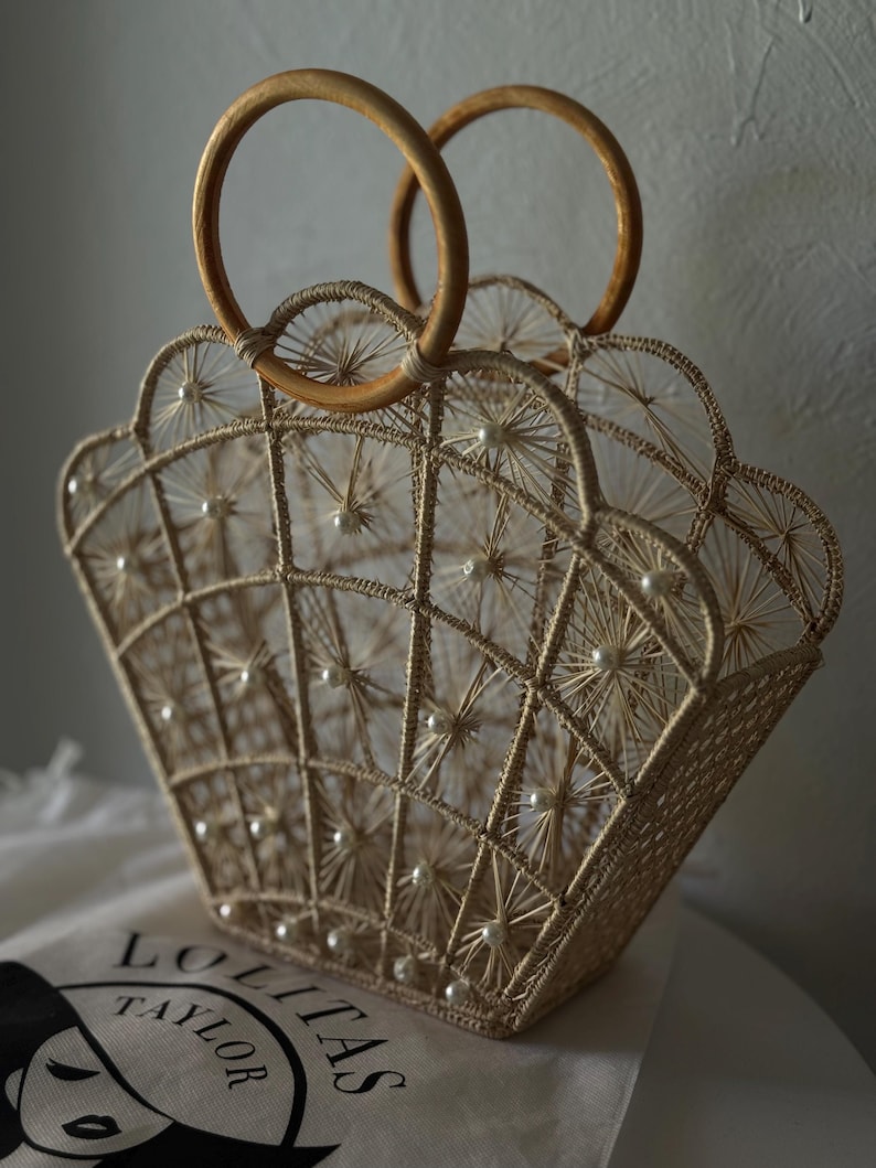 May include: A beige, woven handbag with a shell-like design, featuring wooden handles. The bag is adorned with small, white pearls and has a geometric pattern. The bag sits on a white surface.