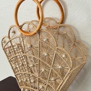 May include: A beige woven handbag with a fan-shaped design, decorated with small white pearls. It has two circular wooden handles. The bag is made of natural fibres and has a geometric pattern.