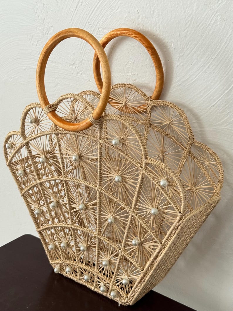 May include: A beige woven handbag with a fan-shaped design, decorated with small white pearls. It has two circular wooden handles. The bag is made of natural fibres and has a geometric pattern.