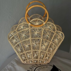 May include: A beige woven handbag with a fan-like design, decorated with small, round embellishments. The bag has two wooden handles and rests on a white cloth bag with a logo.