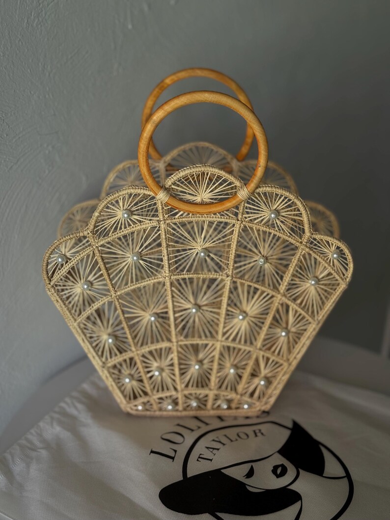 May include: A beige woven handbag with a fan-like design, decorated with small, round embellishments. The bag has two wooden handles and rests on a white cloth bag with a logo.