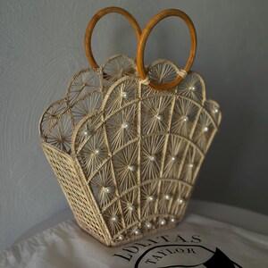 May include: A beige woven handbag with a fan-shaped top and wooden handles. The bag features white pearl-like beads and a geometric pattern. The bag rests on a white cloth with the text "LOLITAS TAYLOR".