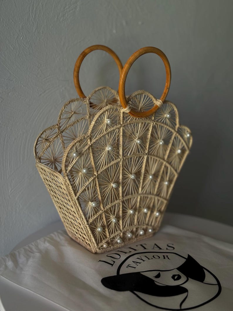May include: A beige woven handbag with a fan-shaped top and wooden handles. The bag features white pearl-like beads and a geometric pattern. The bag rests on a white cloth with the text "LOLITAS TAYLOR".