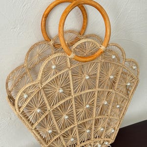 Puede incluir: Un bolso de mano tejido beige con un diseño en forma de abanico. El bolso tiene asas de madera y está adornado con pequeñas cuentas blancas con forma de perla. El patrón tejido crea un diseño geométrico, perfecto para un look de playa o verano.