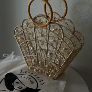 May include: A beige woven handbag with a scalloped edge and wooden handles. The bag has a grid pattern with pearl accents. A white bag with the text "LOLITAS TAYLOR" is visible.