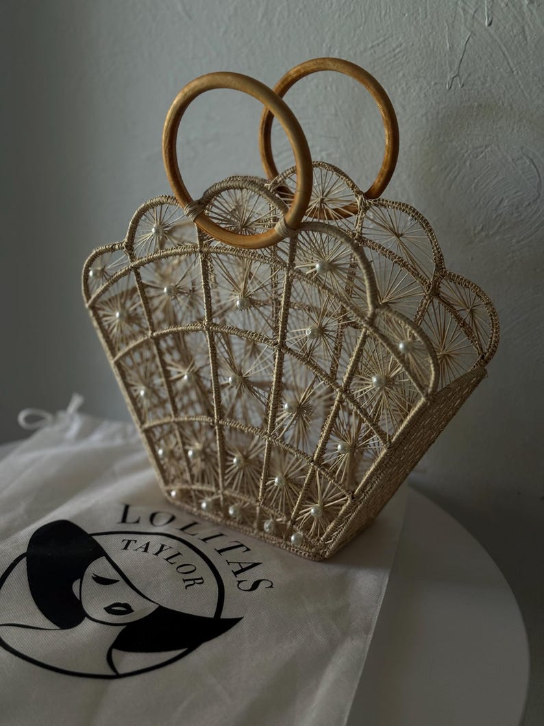 May include: A beige woven handbag with a scalloped edge and wooden handles. The bag has a grid pattern with pearl accents. A white bag with the text "LOLITAS TAYLOR" is visible.