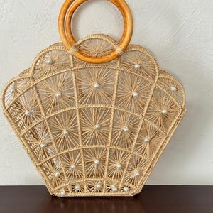 May include: A beige woven handbag with a fan-shaped design, decorated with small white pearl accents. The bag has a wooden handle and an open weave pattern. Ideal for a beach or summer occasion.