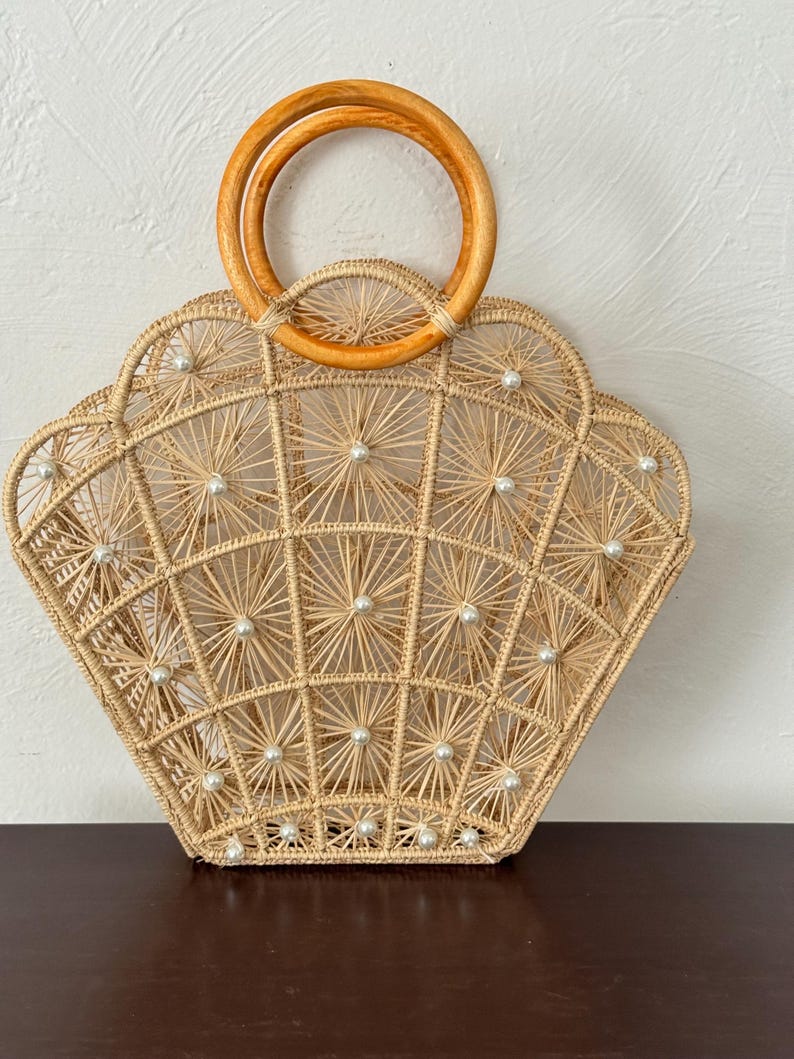 May include: A beige woven handbag with a fan-shaped design, decorated with small white pearl accents. The bag has a wooden handle and an open weave pattern. Ideal for a beach or summer occasion.