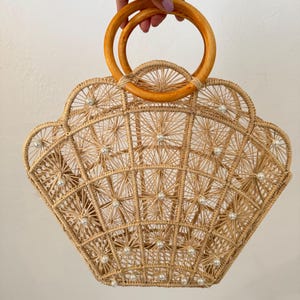 May include: A light brown, woven handbag in a shell shape. The bag has a geometric pattern with small, pearl-like details and wooden handles. The bag is held up by a hand.