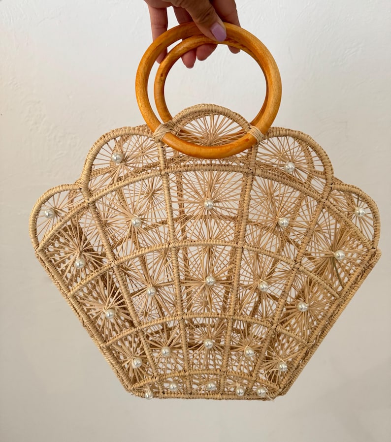 May include: A light brown, woven handbag in a shell shape. The bag has a geometric pattern with small, pearl-like details and wooden handles. The bag is held up by a hand.