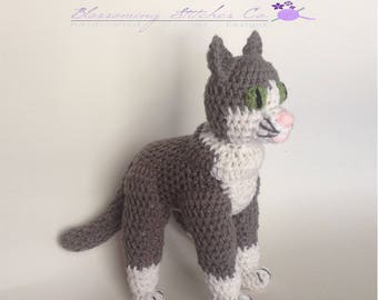 May include: Gray and white crocheted cat toy with green eyes. The cat is standing on a white surface.