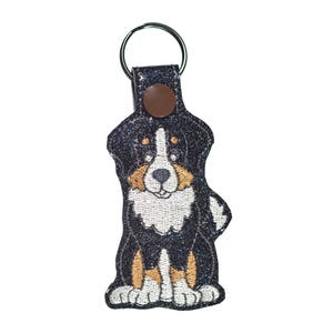 May include: A black, white, and brown Bernese Mountain Dog keychain with a silver key ring. The dog is embroidered on a black fabric with a brown button.