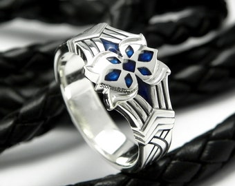 May include: A silver ring with a detailed design featuring a blue cross-shaped inlay. The ring has a geometric pattern and the text "Stones&Fire" is visible. The ring is displayed on a black leather surface.