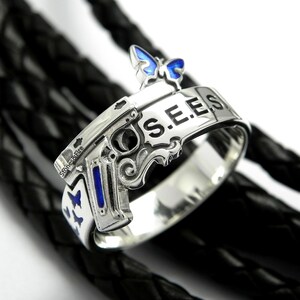 May include: A silver ring featuring a gun design with a blue butterfly on top. The ring is inscribed with "S.E.E.S." on the band.
