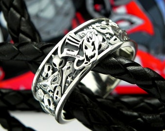 May include: A silver ring with a detailed design featuring a variety of symbols, including a spaceship, an alien, and a star. The ring is on a black braided leather strap.