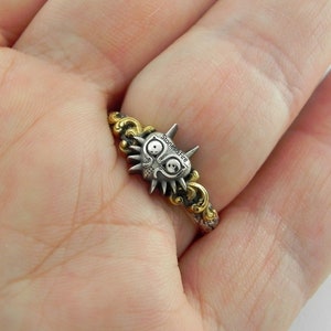May include: A silver and gold ring featuring a stylized mask design. The mask has a detailed face with large eyes and spiky details. The ring band has a decorative gold pattern. The text "stones&fire" is visible on the mask.