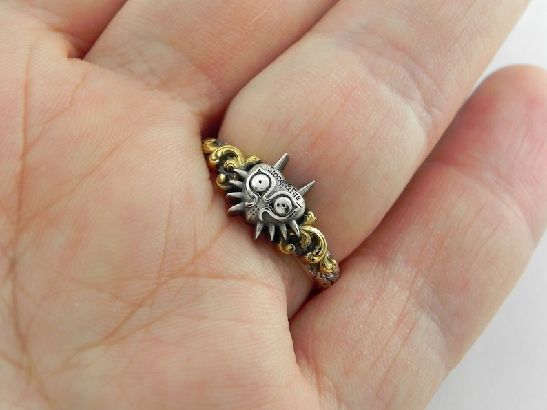May include: A silver and gold ring featuring a stylized mask design. The mask has a detailed face with large eyes and spiky details. The ring band has a decorative gold pattern. The text "stones&fire" is visible on the mask.