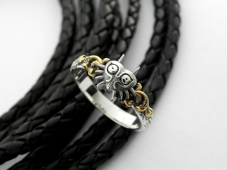 May include: A silver ring with a detailed Majora's Mask design, accented with gold-colored filigree. The ring is set against a black leather background, with the text "Stones & Fire" visible on the ring.
