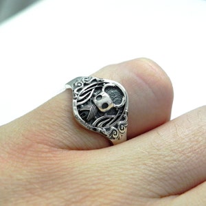 May include: A silver ring with a detailed design featuring a skull and wings.