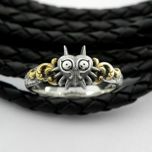 May include: A silver ring featuring a detailed, stylized mask design with gold accents. The ring has a unique, fantasy-inspired aesthetic, with intricate detailing and a polished finish. The ring is set against a black, textured background.