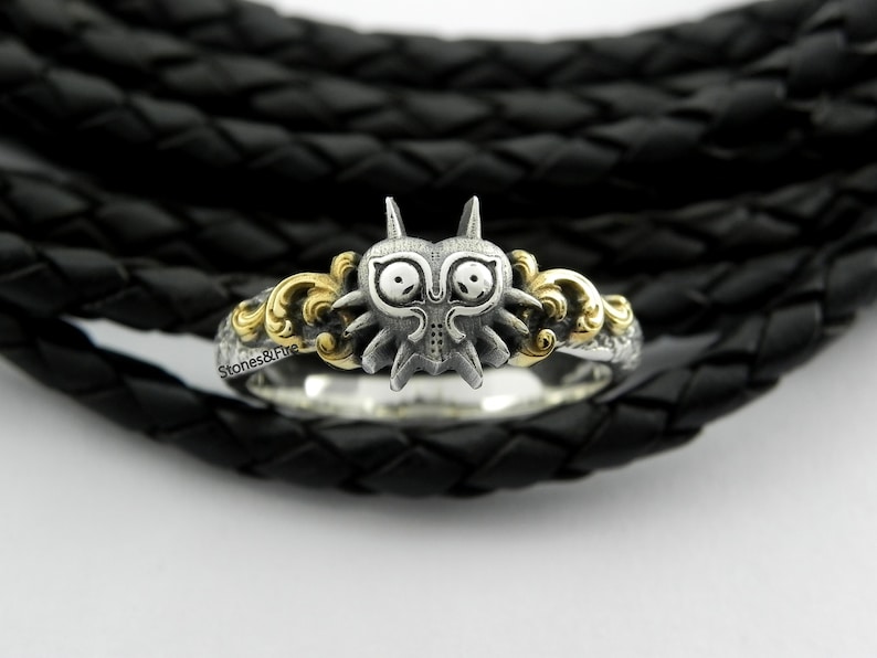 May include: A silver ring featuring a detailed, stylized mask design with gold accents. The ring has a unique, fantasy-inspired aesthetic, with intricate detailing and a polished finish. The ring is set against a black, textured background.