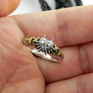 May include: A silver ring featuring a detailed, stylized mask design with gold accents. The ring has a unique, fantasy-inspired aesthetic, with intricate detailing and a two-tone color scheme. The ring is held in a hand.