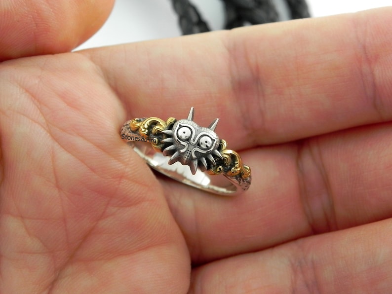May include: A silver ring featuring a detailed, stylized mask design with gold accents. The ring has a unique, fantasy-inspired aesthetic, with intricate detailing and a two-tone color scheme. The ring is held in a hand.