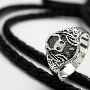 May include: A silver ring with a detailed design of a knight with a helmet and wings. The ring is on a black braided leather background.