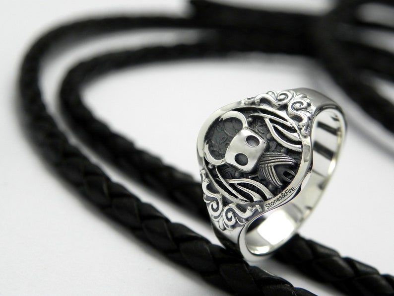 May include: A silver ring with a detailed design of a knight with a helmet and wings. The ring is on a black braided leather background.