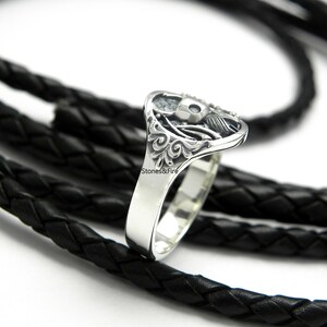 May include: A silver ring with a detailed skull design and intricate floral accents. The ring is on a black braided leather cord.