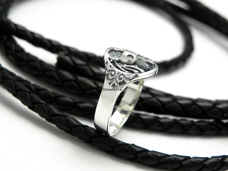 May include: A silver ring with a detailed skull design and intricate floral accents. The ring is on a black braided leather cord.