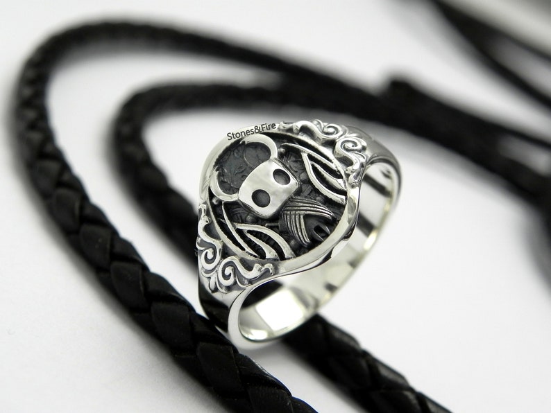 May include: A silver ring with a detailed design of a knight's helmet with wings and a floral border. The ring is on a black braided leather background.
