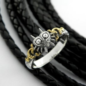 May include: A silver ring with a detailed Majora's Mask design, featuring a stylized face with horns and eye details. The ring has gold accents and is set against a black background.