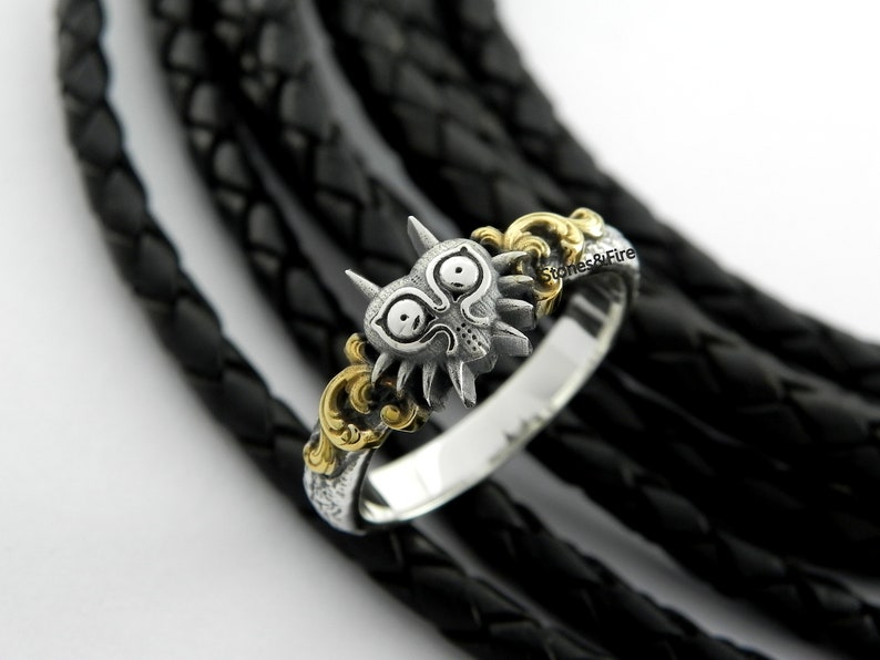 May include: A silver ring with a detailed Majora's Mask design, featuring a stylized face with horns and eye details. The ring has gold accents and is set against a black background.