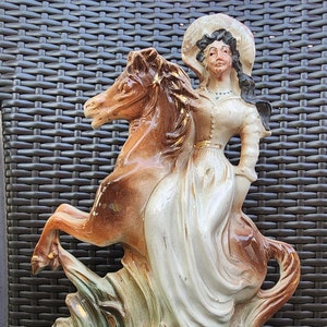 May include: A ceramic figurine depicting a woman riding a rearing horse. The sculpture features a woman in a white dress and hat, with the horse in shades of brown. The base is teal with gold accents. The figurine is approximately 12 inches tall.