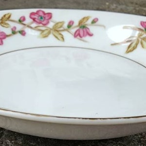 May include: White oval-shaped porcelain bowl with a gold rim and floral design. The bowl features pink flowers with blue centers and gold-colored leaves. The floral pattern decorates the rim of the bowl.