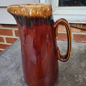 May include: A brown and tan glazed ceramic pitcher with a wide mouth and a handle. The pitcher has a brown glaze with a tan glaze on the top rim.