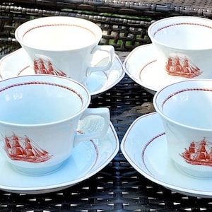 May include: Set of eight white teacups and saucers with a red trim and a red and brown ship design.