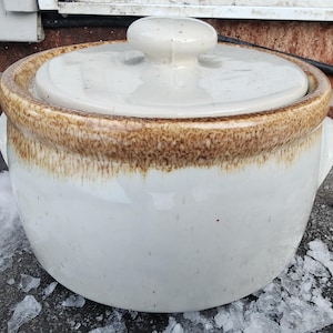 May include: A white ceramic casserole dish with a lid. The rim and lid edge have a brown, speckled glaze. The dish has two side handles and a knob on the lid. The dish is sitting on a surface with ice.