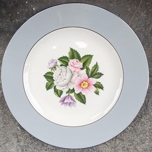May include: A round, light blue plate with a floral design. The center of the plate features a bouquet of white, pink, and purple flowers with green leaves. The plate has a light blue border.