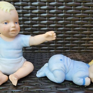 May include: Two porcelain baby figurines, one sitting and one lying down. Both are wearing blue and white clothing and have blonde hair.