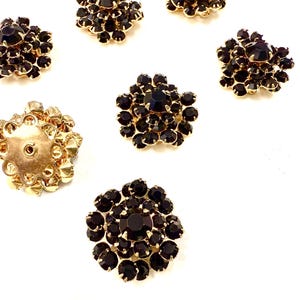 May include: A collection of vintage black rhinestone buttons with gold-tone metal settings. The buttons are arranged on a white surface, showcasing their intricate floral designs and sparkling facets. One button is shown from the back.