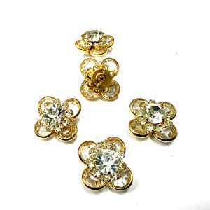 Può includere: Cinque abbellimenti a forma di fiore con pietre centrali chiare e sfaccettate e montature in metallo dorato. Gli abbellimenti sono disposti su uno sfondo bianco, mostrando i loro dettagli scintillanti.