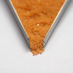 Op de afbeelding: Een metalen bak gevuld met kleine, oranje kralen. De kralen vallen uit de bak op een wit oppervlak.