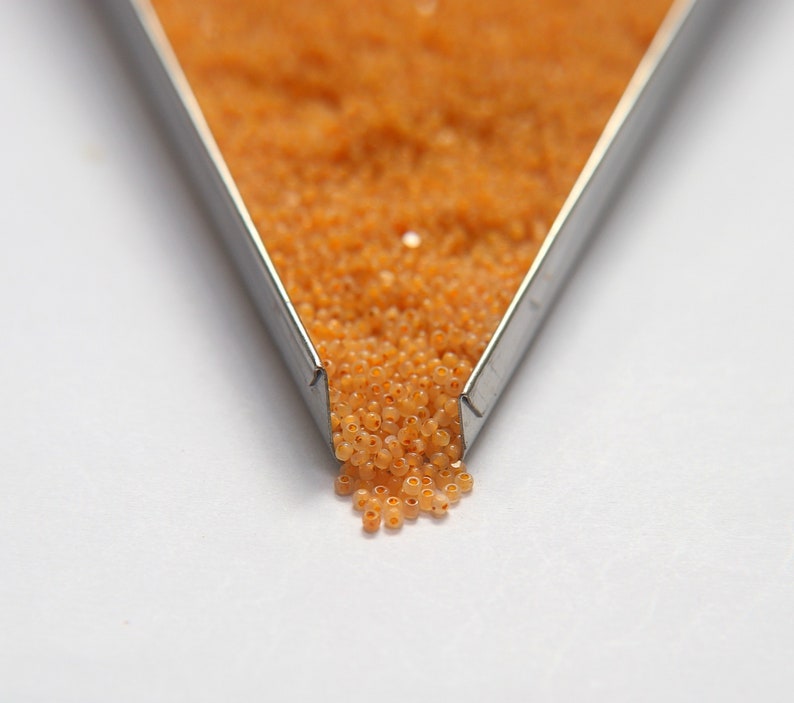 Op de afbeelding: Een metalen bak gevuld met kleine, oranje kralen. De kralen vallen uit de bak op een wit oppervlak.
