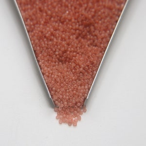 Peut inclure: Gros plan d'une cuill&egrave;re en m&eacute;tal remplie de petites perles de rocaille rose clair. Les perles se r&eacute;pandent de la cuill&egrave;re sur une surface blanche.