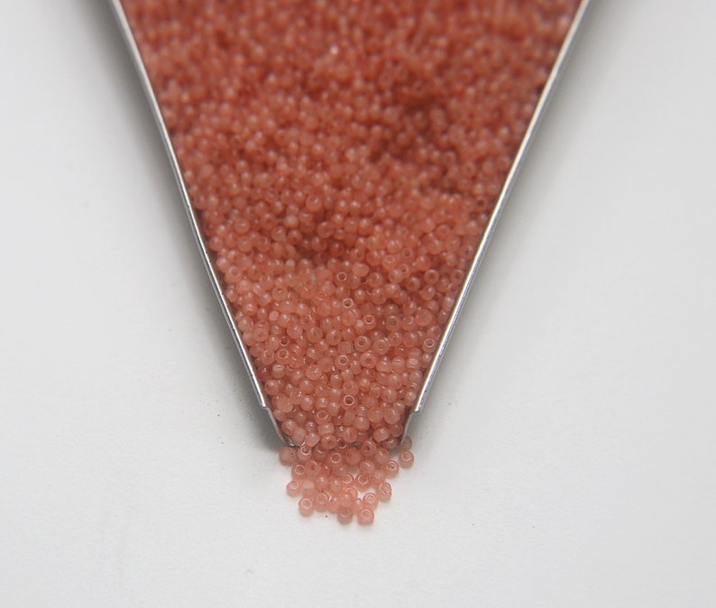 Peut inclure: Gros plan d'une cuill&egrave;re en m&eacute;tal remplie de petites perles de rocaille rose clair. Les perles se r&eacute;pandent de la cuill&egrave;re sur une surface blanche.