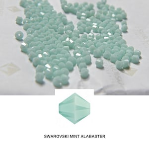 Puede incluir: Primer plano de un montón de pequeñas cuentas de vidrio facetadas de color azul claro. Las cuentas están dispuestas en un patrón aleatorio y todas son del mismo tamaño. Las cuentas están hechas por Swarovski y se llaman "Mint Alabaster".
