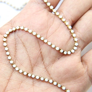 Puede incluir: Un delicado collar de cadena dorada con peque&ntilde;as piedras preciosas blancas y redondas. Las piedras est&aacute;n espaciadas uniformemente a lo largo de la cadena, creando un aspecto brillante y elegante. El collar est&aacute; sobre una mano.
