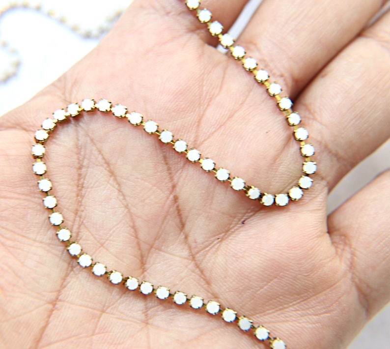 Puede incluir: Un delicado collar de cadena dorada con peque&ntilde;as piedras preciosas blancas y redondas. Las piedras est&aacute;n espaciadas uniformemente a lo largo de la cadena, creando un aspecto brillante y elegante. El collar est&aacute; sobre una mano.